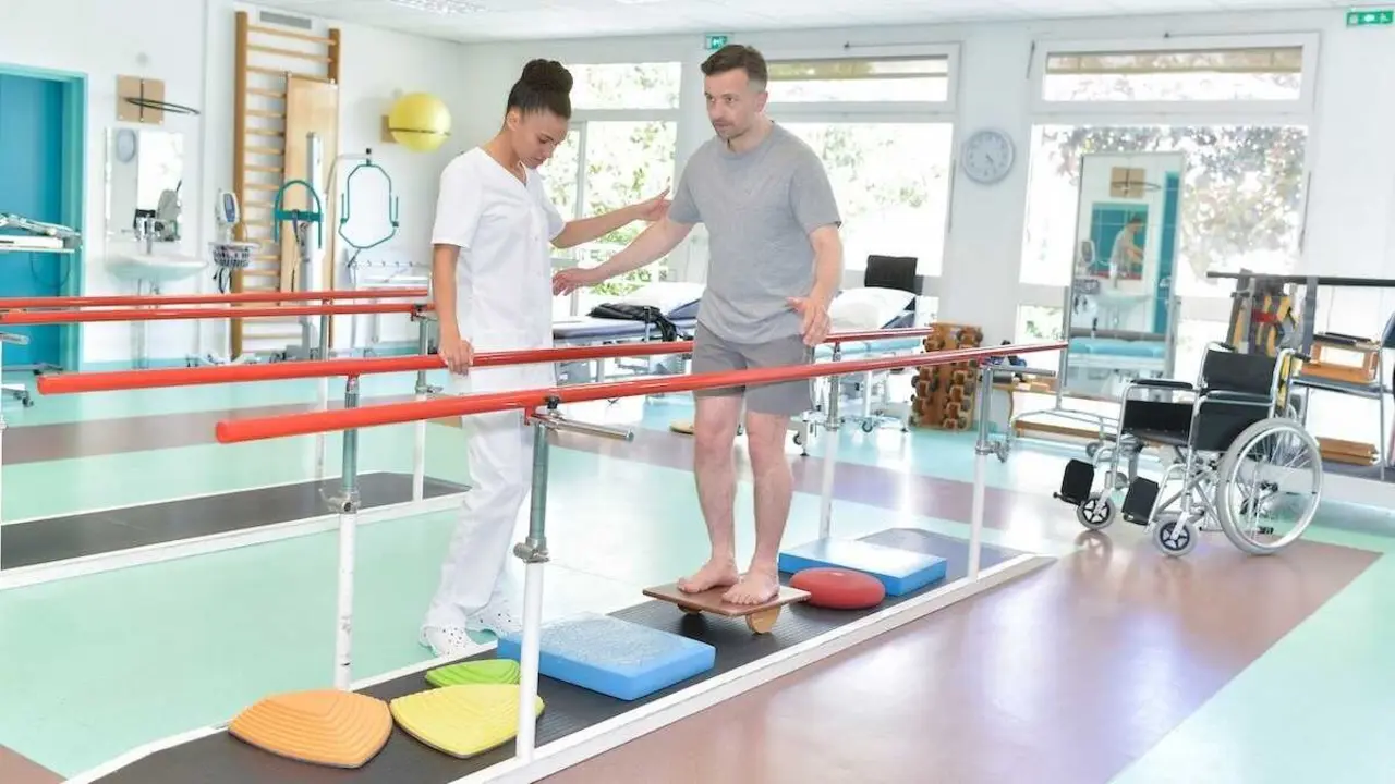 Un fisioterapeuta supervisa a un paciente durante un ejercicio de equilibrio sobre tabla basculante en una sala de rehabilitaci&oacute;n neurol&oacute;gica.